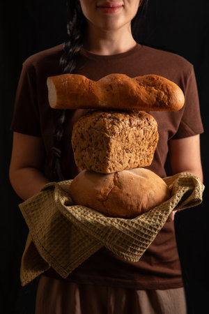 brunette girl with a braid holds a stack of freshly baked breads in her hands. She wears a brown t shirt standing in a warm indoor space showcasing her love for baking and fresh goods.の写真素材