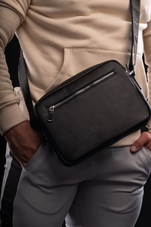 man dressed in a beige hoodie showcases a black bag emphasizing his casual style. He stands in front...