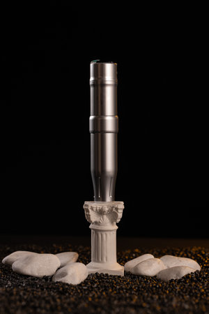 A tattoo machine stands upright on a detailed stone base surrounded by white pebbles. The dark...