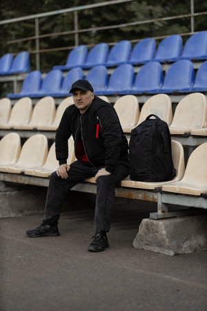 man dressed in a black bomber jacket and cap sits quietly in an empty stadium tribune. He has a...