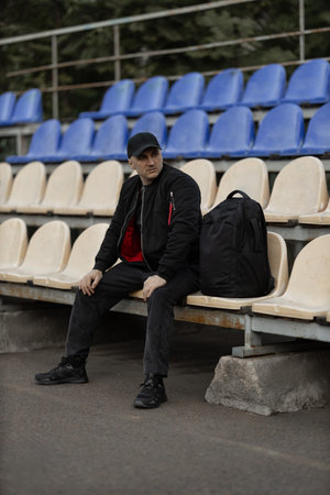 man wearing a black bomber jacket and cap sits on a bench in an empty stadium tribune. He has a backpack beside him and appears to be in a reflective mood in a quiet setting.の写真素材