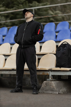 Man wearing black bomber jacket and cap stands in empty bleachers displaying emotions as he watches...