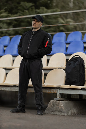 man wearing a black bomber jacket and cap stands in the empty stands focused on the game below him. His body language shows anticipation and engagement with the teams performance.の写真素材