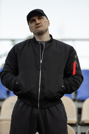 In an empty stadium man wearing black bomber jacket and cap stands with arms crossed expressing...