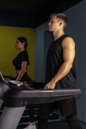 sporty girl and a man wearing headphones walk side by side on treadmills focusing on their fitness...