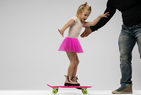 small blonde girl is learning to skateboard while wearing a pink tutu. An adult is helping her maintain balance during the activity.の写真素材