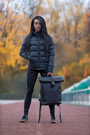 Brunette woman in black jacket and black backpack stands confidently on a track. Trees in the background show colorful leaves. It is a sunny day in fall season.の写真素材