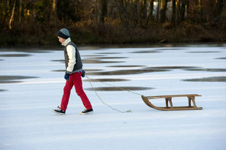 A boy is walking on a frozen pond with his sledgeの写真素材
