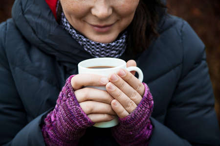 White Cup with tea in hand. Knitted purple fingerless gloves women's hands. Autumn photography.の写真素材