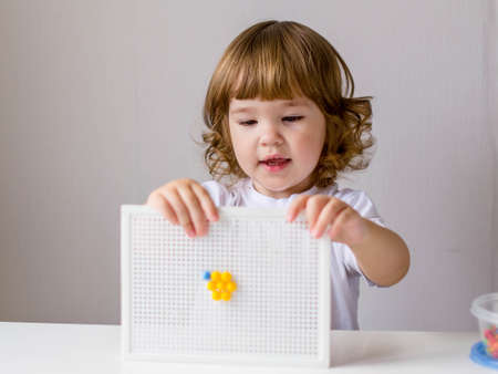 A little curly-haired girl in a white t-shirt plays at a white table in a multicolored plastic mosaic. The child shows the result of his creativity. The concept of early education, the development of fine motor skills.の写真素材