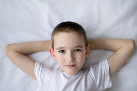 Top view of a boy who is lying in bed and folded his arms behind his head. The child lies on his back on a white background.の写真素材