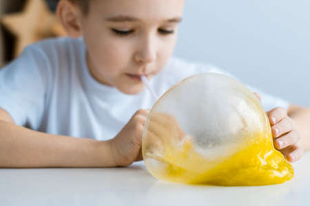 A boy in a white T-shirt at a white table plays a slime. Child inflates bubble slime.