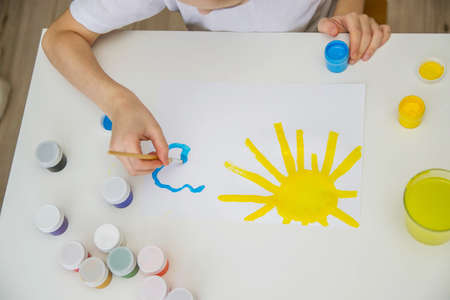 A child sits at a white table and draws clouds. Children's drawing colorful.の写真素材