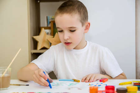 A boy in a white T-shirt sits at a white table and draws with pencils and paints.の写真素材