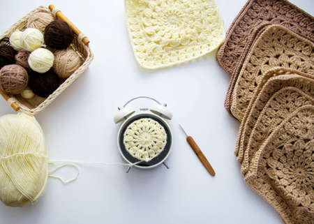 Crochet on a white background. On a white table are balls of yarn, square motifs, an alarm clock, a...