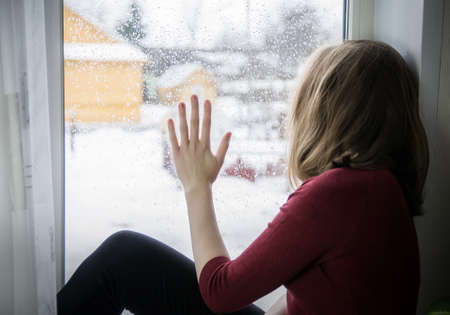 A teenager girl sits on a windowsill and looks out the window, holds one hand on the glass. The...