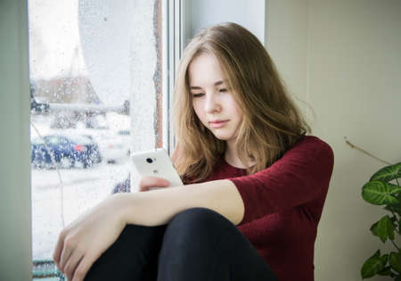 Overweight teenager girl with long blond hair is sitting by the window on the windowsill with a...