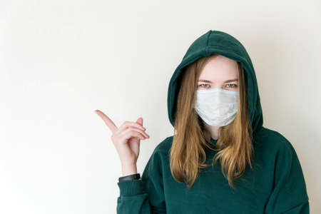 A girl in a medical mask, in a green sweatshirt in a hood points to a place for text. Banner for quarantine, self-isolation, coronavirus pandemic.の写真素材