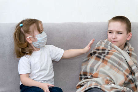 Keep a distance with sick people. A boy and a girl in a mask sit on a sofa in the distance and the girl pushes away the sick boy.の写真素材