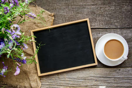 Top view of a black slate board in a wooden frame, a white cup with a saucer and coffee with milk, a bouquet of wild flowers on an old rustic wooden table.の写真素材