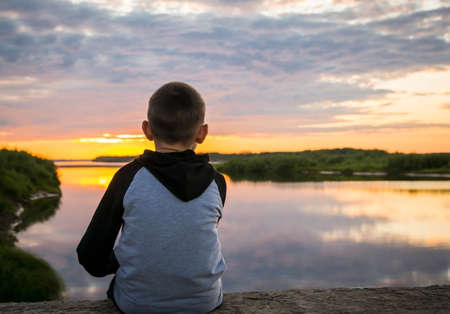 sad, lonely child sits alone by the river at sunset, rear viewの写真素材