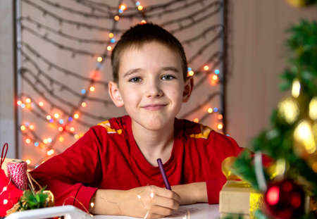 A boy in a red jacket writes a letter to Santa Claus at the table.