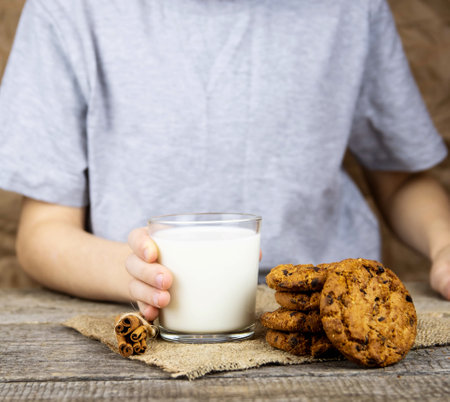 The child sits at a wooden table and holds a glass of milk, oatmeal cookies on the table.の写真素材