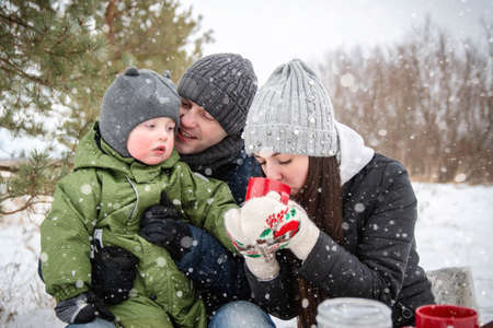 The family in winter in the park on a picnic. Mom, dad and baby sit at the table and look right into...