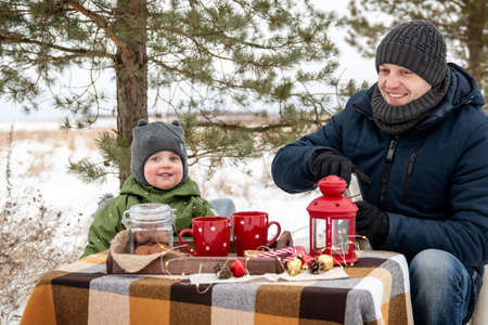 Winter picnic in the woods. Dad and son sit at the table and drink hot tea from the flaskの写真素材