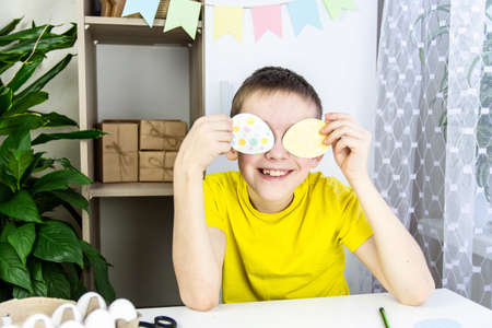 The concept process of preparing for Easter. A boy in a yellow T-shirt holds paper eggs in his eyes...