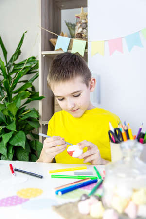 A boy in a yellow T-shirt sits at a table and paints a white egg with colored markers. The concept...