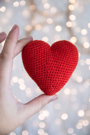 Knitted red heart in the hand against the background of a garland. The concept of love and...