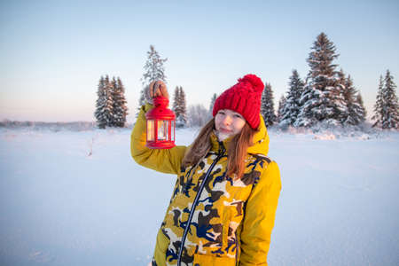 A teenage girl joyfully holds in her hands a red candlestick lantern in the winter forest.の写真素材