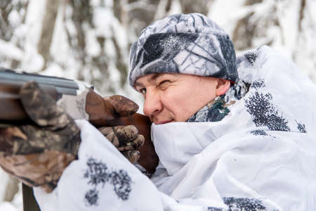 The concept of winter hunting. A male hunter in a white camouflage suit shoots a gun.
