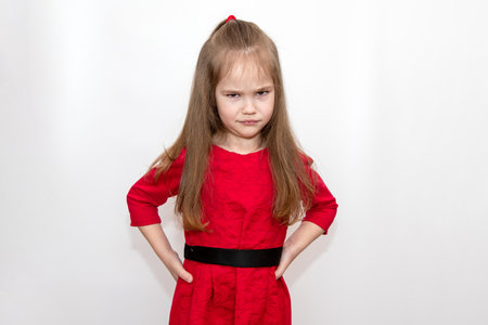 Portrait of a girl in a red dress with long hair on a white background. The child stands frowning,...