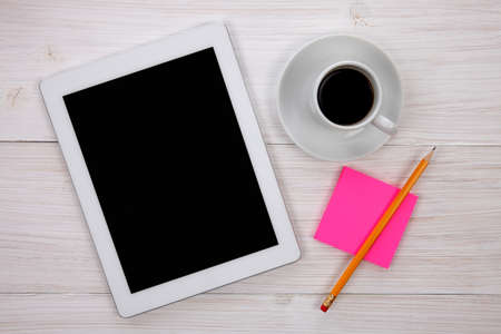 Workplace with hands holding blank digital tablet, pink notepad with yellow pencil and cup of coffee on wooden background. Above view shot.の写真素材