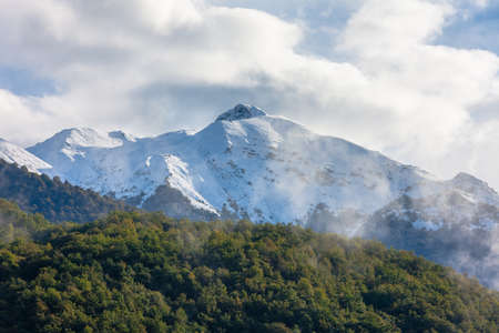Mountain peak covered with snow, green forest, clouds
