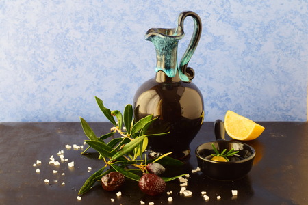 Traditional Greek ceramic olive oil jar with lemon, sea salt, olives, brunch of olive tree on a dark back ground.の写真素材