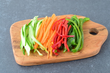 Fresh cucumber, carrot, red and green sweet paprika sliced in stripes on an olive wood cutting board on a grey abstract background. Step by step cookingの写真素材