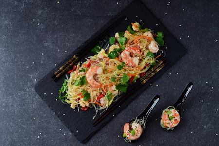 Noodles with prawns and vegetables on a black stone plate and in traditional soup spoons on a grey abstract background. Healthy food conceptの写真素材