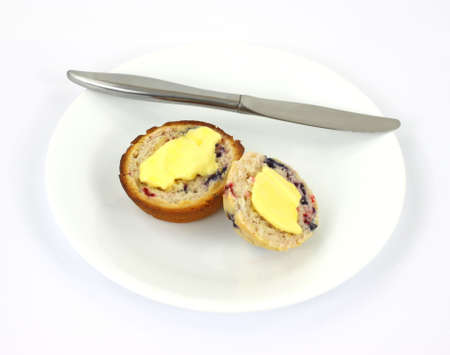 A freshly baked buttered berry muffin on a white plate with a knife against a white background.の写真素材