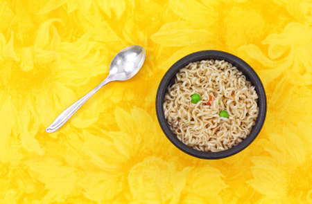A serving of noodles peas and carrots in a black bowl with spoon on a yellow floral cloth background.の写真素材
