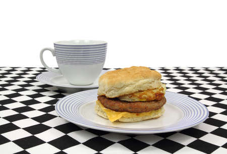A freshly made sausage egg and cheese breakfast sandwich on a blue striped plate with coffee cup on a black and white checkerboard cloth.の写真素材