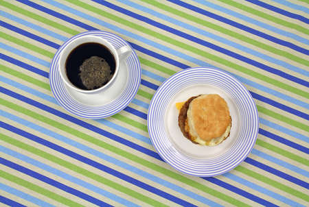 A freshly made sausage egg and cheese breakfast sandwich on a blue striped plate with black coffee.の写真素材