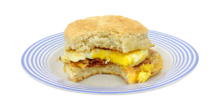 A freshly made breakfast sandwich biscuit with bacon egg and cheese that has been bitten once on a blue striped plate.の写真素材