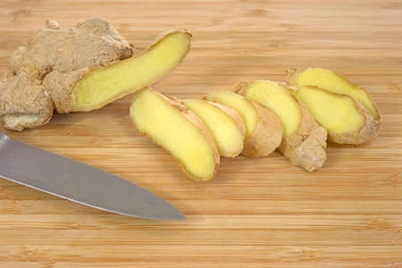 Close view of a ginger root with several slices and the blade of a knife on a wood cutting board の写真素材