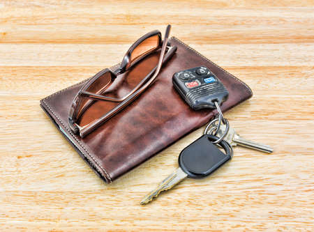 A set of car keys and tinted sunglasses atop a brown leather wallet on wood tabletop 