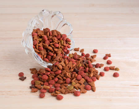 A glass bowl tipped on side with generic dry cat food spilling onto a wood counter top.の写真素材