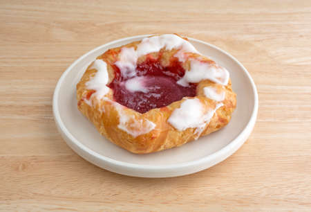 A single raspberry filled danish pastry on a small plate atop a wood table top illuminated with natural light.の写真素材