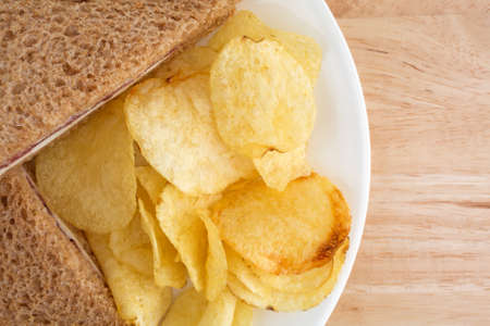 Top close view of a roast beef wheat bread sandwich with white cheese and mayonnaise plus potato chips on a white plate atop a wood table top.の写真素材